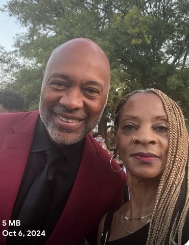 A smiling bald man with a short gray beard in a maroon suit jacket beside a woman with long blonde braids and a nose ring, outdoors among green trees