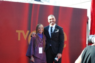 A woman in a purple dress and a man in a dark suit smiling together in front of a large red television backdrop