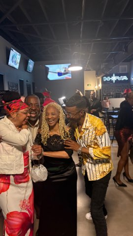 Four friends laughing together in a dimly lit lounge bar, a neon sign glowing on the wall behind them