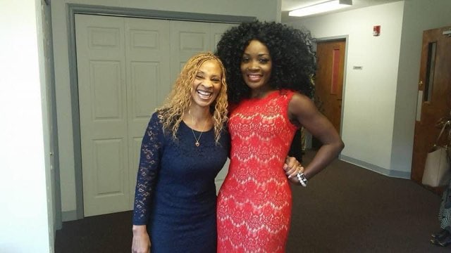 Two women together in a bright hallway, one in a long-sleeved dark blue lace dress and one in a sleeveless red lace dress