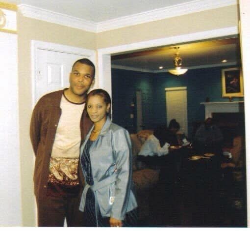 A smiling man in a brown jacket and a woman in a light blue jacket standing together indoors, an open doorway behind them