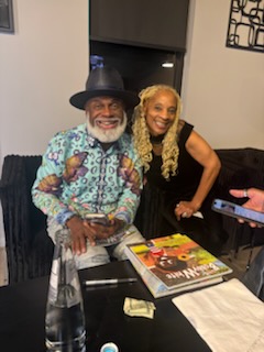 A gentleman with a white beard and a black hat beside a woman with long braids, magazines on the table in front of them