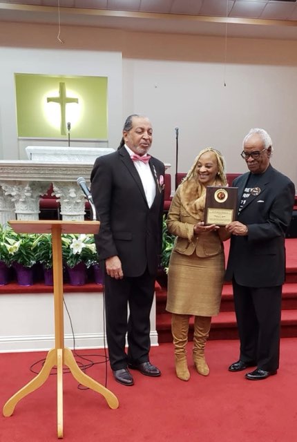 Three people on the red carpet of a church in front of the altar — a man in a black suit and pink bow tie, a woman in gold holding a wooden plaque, and a man in a black suit and glasses