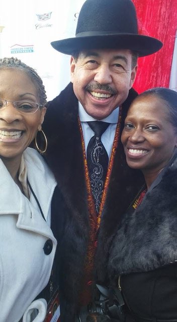 Three people on a red carpet — a gentleman in a black hat and fur-trimmed coat between two smiling women