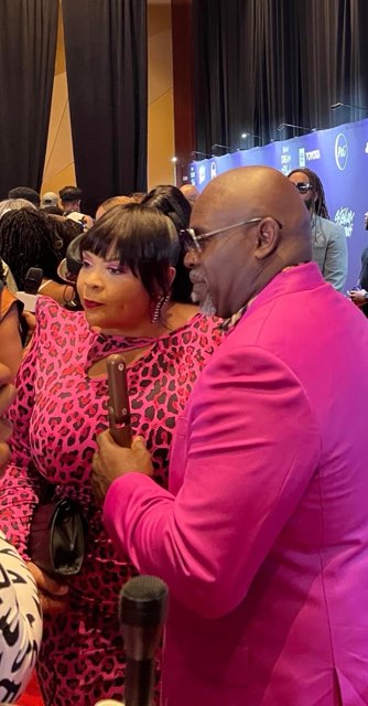A smiling man in a bright pink suit and glasses holding the microphone beside a woman in a pink and black leopard-print dress, a branded blue backdrop behind them
