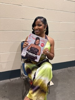 A smiling woman sitting on a folding chair holding up a magazine cover with her photograph on it