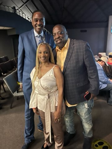 Three people together in an auditorium — two men in blazers either side of a smiling woman with blonde braids