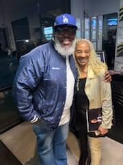 A tall smiling man with a thick white beard in a blue Dodgers cap and bomber jacket, arm in arm with a woman in a metallic gold jacket, in a warmly lit restaurant