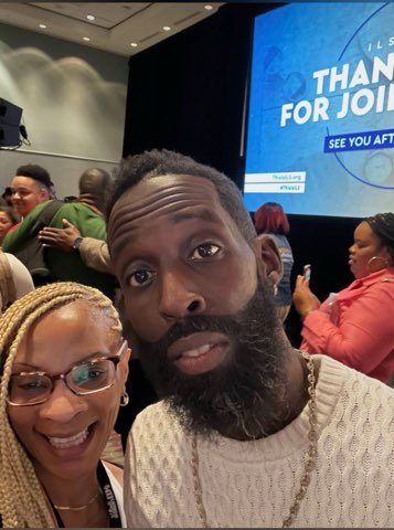 Two smiling people together at a large event hall — a woman with glasses and braided hair beside a man in a white textured sweater, a big blue screen behind them