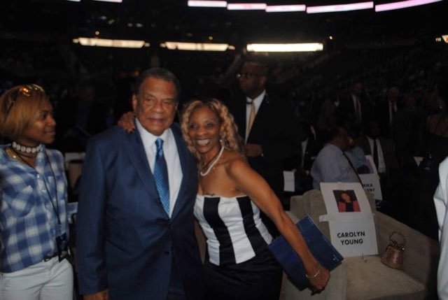 A joyful moment between a man in a sharp blue suit and a woman in a black and white striped dress at a large indoor event, a name card on a chair in the foreground