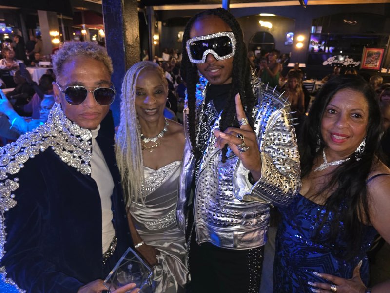 A celebratory group of four in bold evening fashion — silver embellishment, a silver dress, a spiked jacket and a blue sequined gown — at a crowded reception