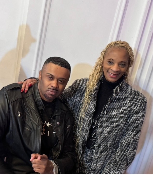 A man in a black leather jacket with a cross necklace beside a woman with long blonde braids in a black and white tweed coat
