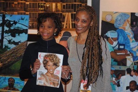 Two smiling readers holding up a magazine together in front of a wall of framed artwork