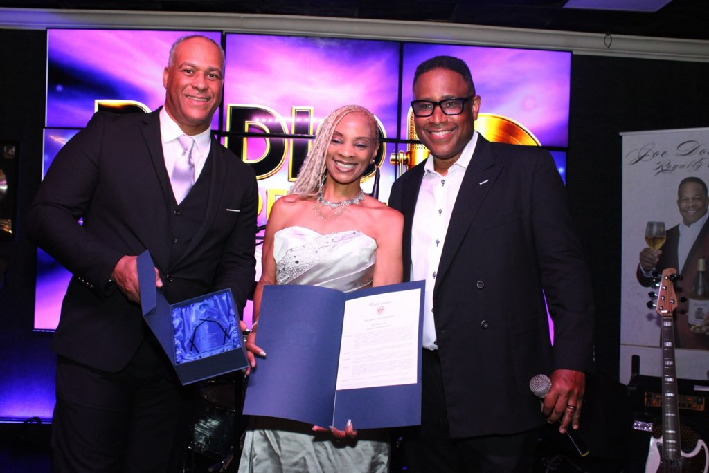 Three people on a stage: a man holding an open blue box, a woman in a white dress holding a certificate, and a man with a microphone