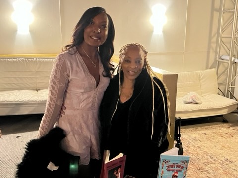 Two smiling women together at a book signing, one in a pale pink dress, the other holding copies of a book titled Chronicles