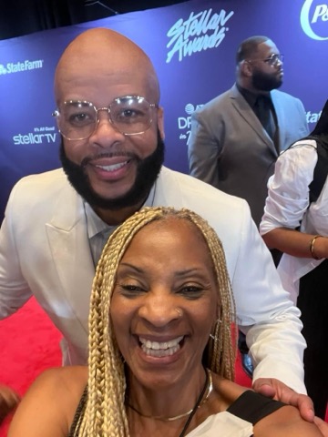 A man in a pale suit jacket and clear glasses standing behind a smiling woman with braided hair on a red carpet, a Stellar Awards and State Farm backdrop behind them