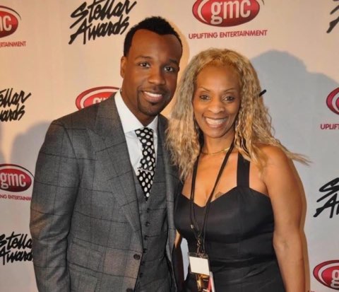 A man in a grey plaid suit and a woman in a black sleeveless top, smiling together in front of a Stellar Awards and gmc Uplifting Entertainment backdrop