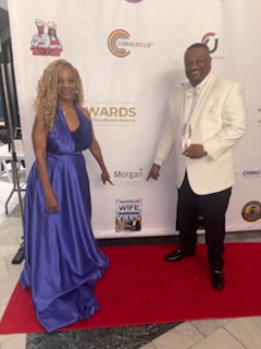 Two elegantly dressed people on a red carpet in front of a branded awards backdrop