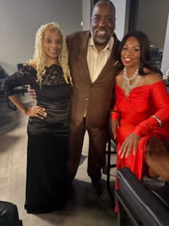 Three people together at an indoor event — a woman in a long black dress, a man in a brown suit, and a woman in a red off-the-shoulder dress