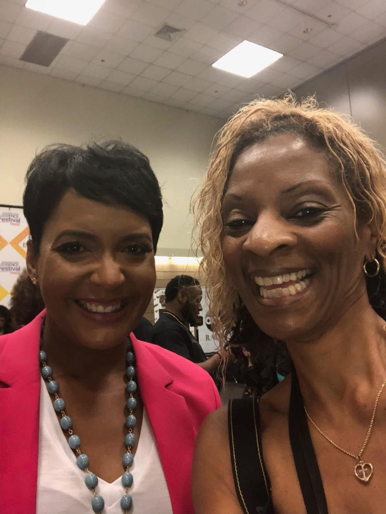 Two smiling women together in front of an Essence Festival backdrop, one in a bright pink blazer and blue beaded necklace