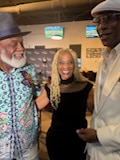 Three people in a bar — a gentleman with a white beard in a grey fedora and blue patterned shirt, a woman with long blonde braids, and a man in a white flat cap and white jacket