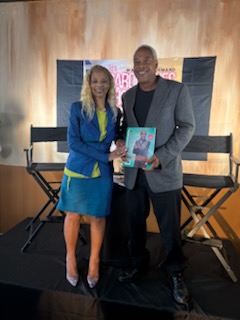 Two smiling professionals on a small stage holding up a book together in front of a university backdrop