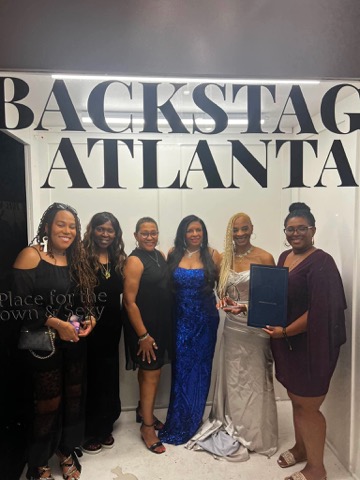 Six women in formal dress together in front of a Backstage Atlanta sign, one holding a certificate