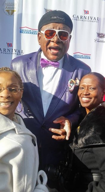 A man in a purple suit and bow tie with two smiling women, in front of a Trumpet Awards and Carnival backdrop