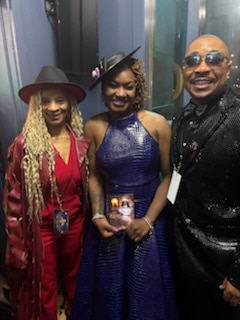 Three elegantly dressed people together — a woman in red with a wide-brimmed hat, a woman in the center in a blue gown holding a small framed photograph, and a woman in a black sequined top and sunglasses