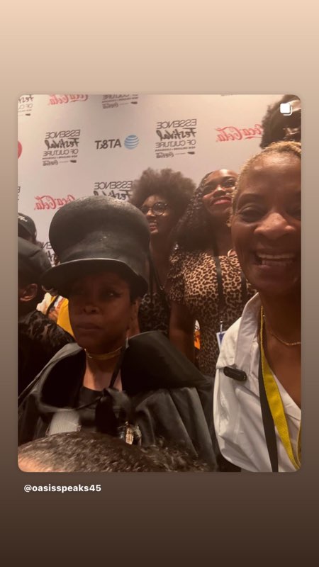 A group photograph at the Essence Festival — a striking figure in a black top hat and cape beside a smiling woman in white, in front of the AT&T backdrop