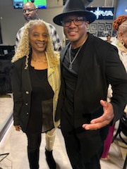 A woman with long blonde curls beside a man in a black hat and glasses, a restaurant sign behind them