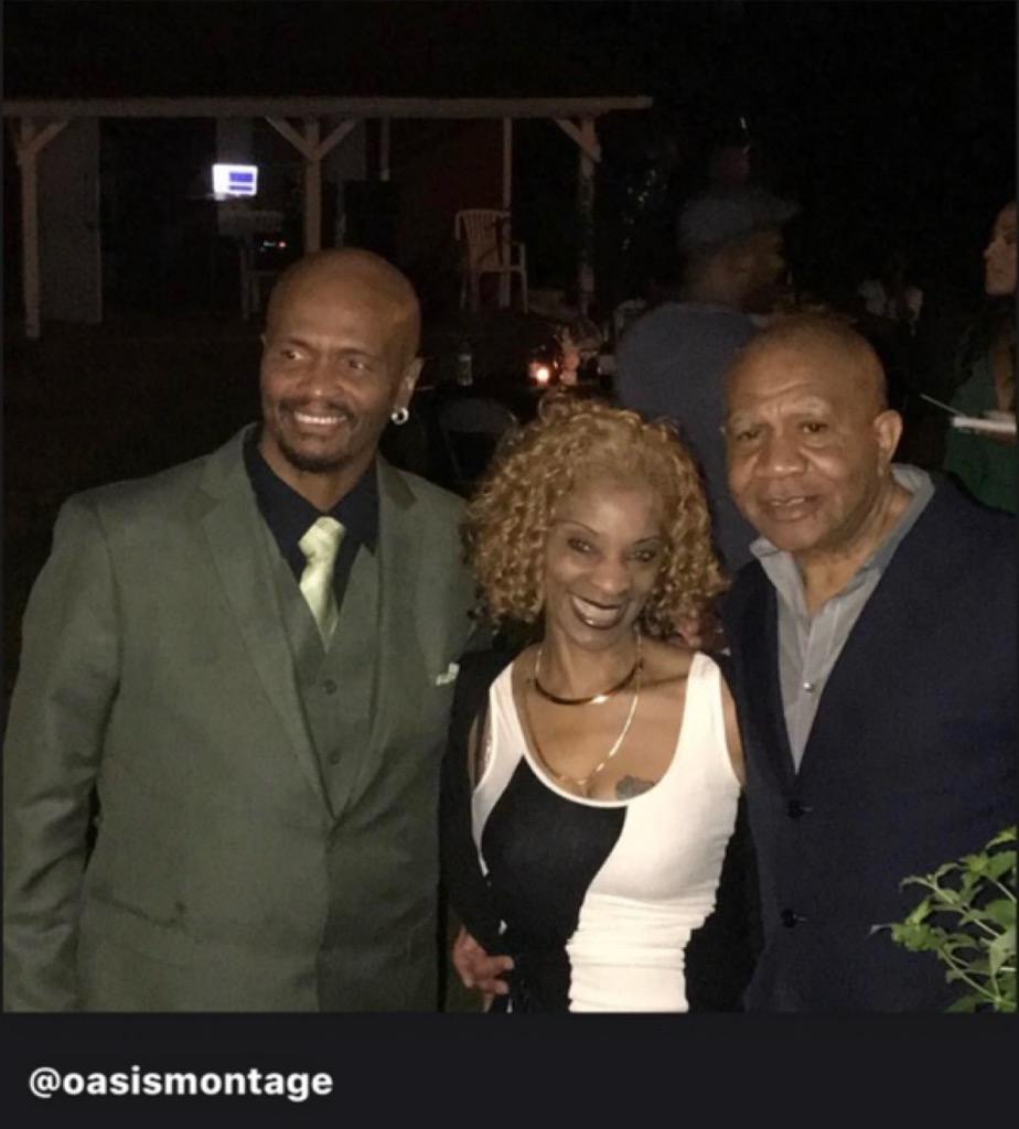 Three smiling people together at an outdoor evening event — a man in a green suit, a woman in a black and white top, and a man in a navy blazer