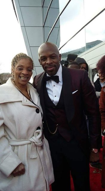 A woman in a white belted coat and a man in a black tuxedo and bow tie on a red carpet outside a modern glass building