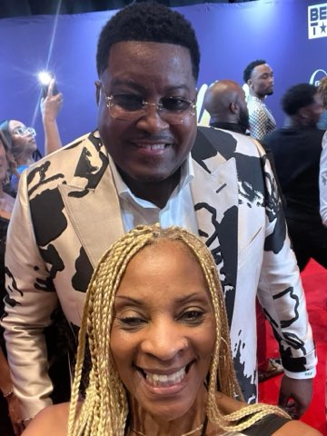 A smiling man in a white jacket patterned with bold black shapes standing just behind a woman with blonde braids at a red carpet event