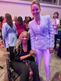 A woman in a lavender blazer standing hand in hand with a smiling woman seated in a wheelchair at an event