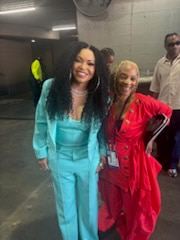 Two smiling women backstage in bright turquoise and red outfits, a lanyard around one neck