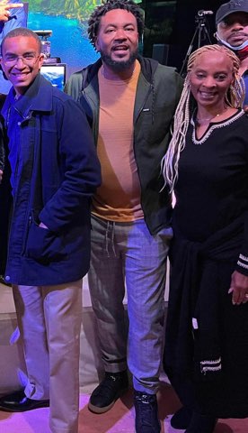 Three smiling people together at a media or production set, a television screen glowing behind them